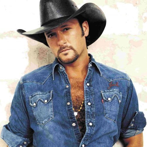 A man in a denim shirt and black cowboy hat stands against a light background, exuding the classic vibe of Tim McGraw. For booking inquiries related to Tim McGraw, please reach out to us.