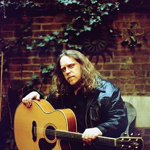 Long-haired musician in a leather jacket sits with an acoustic guitar, evoking vibes reminiscent of Warren Haynes, against a brick wall adorned with ivy.